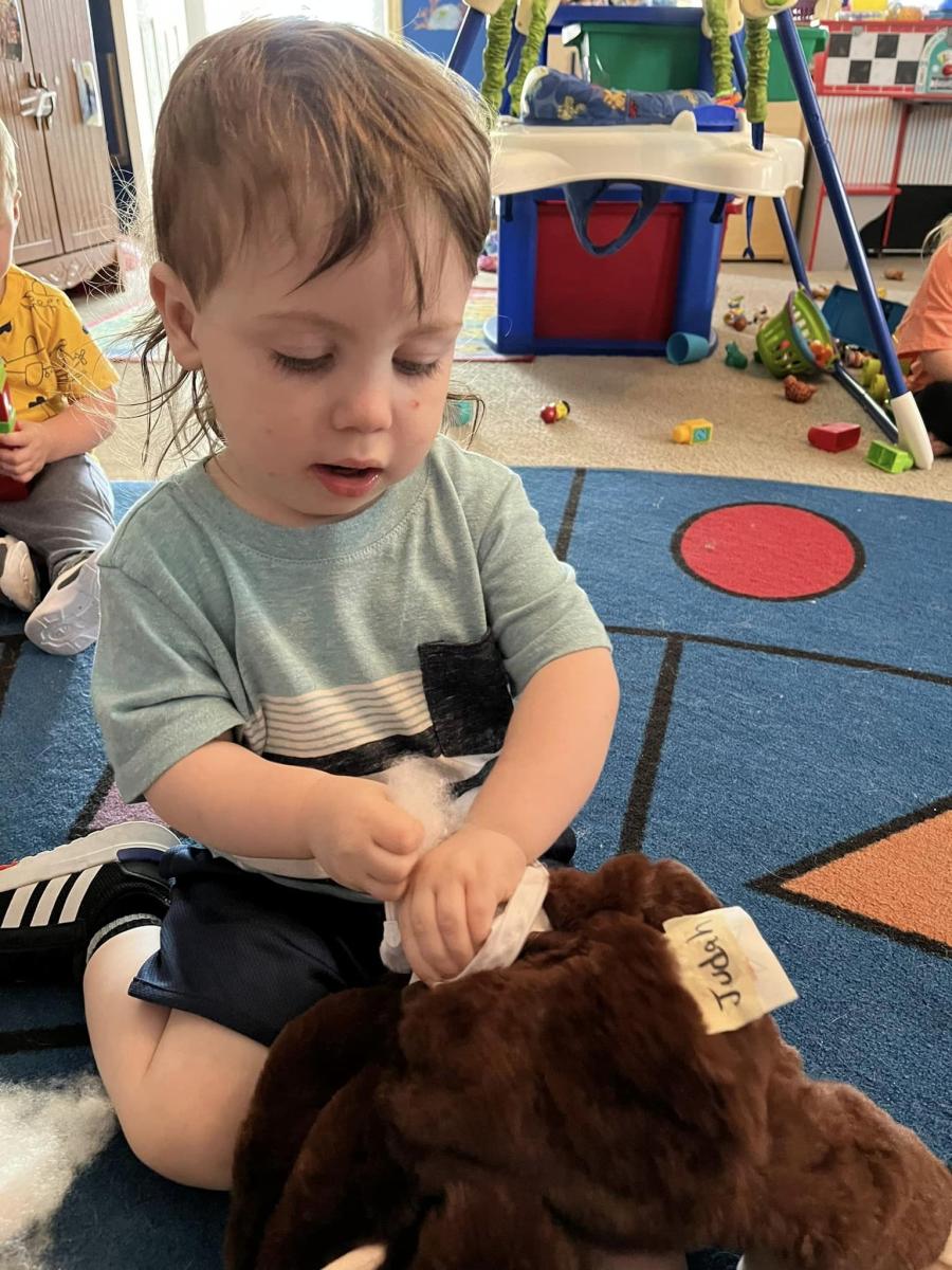 A toddler sitting on a play rug, stuffing a brown teddy bear